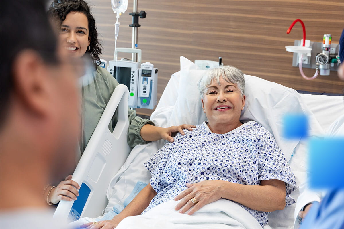 Elderly patient lies in a hospital bed, smiling as a visitor touches her shoulder; IV pump and medical equipment visible behind.