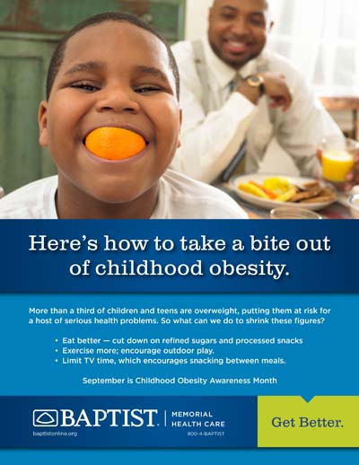 Boy holds an orange slice in his mouth, smiling while a man sits at a breakfast table behind him.