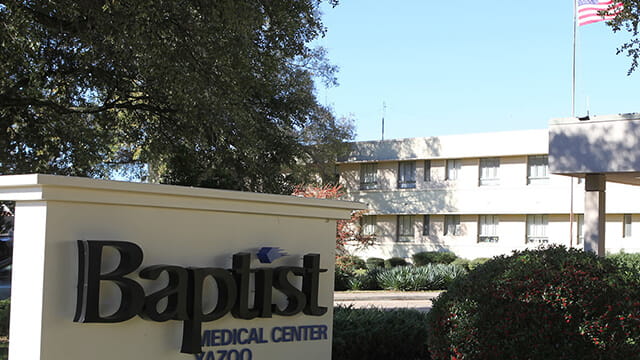 Exterior view of Baptist Medical Center Yazoo, showing the entrance canopy, landscaped grounds, and the hospital building with an American flag flying above.