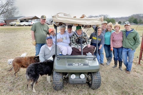 Parked "Club Car" golf cart with people posing around it, smiling in a grassy rural field; two dogs stand nearby.