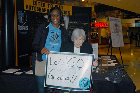Two women hold a whiteboard cheering in an arena concourse with Memphis Grizzlies signage.