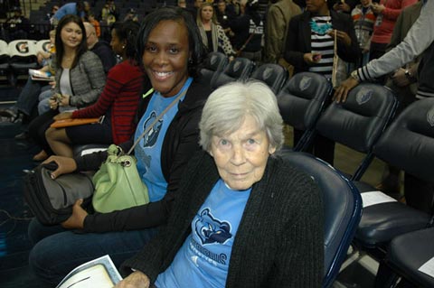 Elderly woman and younger woman seated courtside at a basketball arena, wearing blue team shirts with a bear logo, smiling.