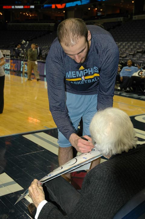 Basketball player signs autograph for a seated elderly fan at an indoor arena. Text in image: "MEMPHIS BASKETBALL"