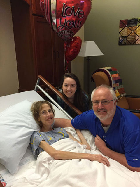 Patient reclines in bed, smiles and holds hands with two visitors; room with lamp, armchair, heart-shaped balloon reading "Love you!".