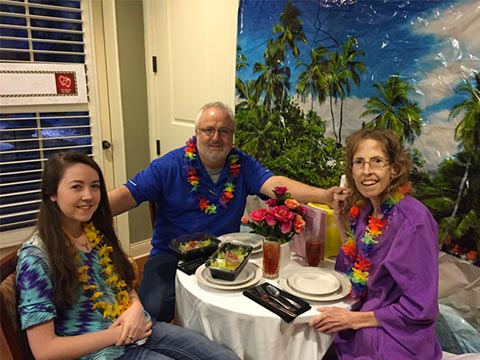 Three people wearing colorful leis sit at a small table eating and smiling, indoors with tropical backdrop and window blinds.
