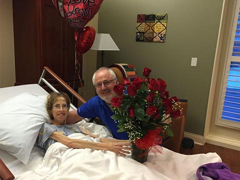 Patient in hospital bed receives large bouquet of red roses from smiling man beside bed, hospital room with balloons reading "Love You".